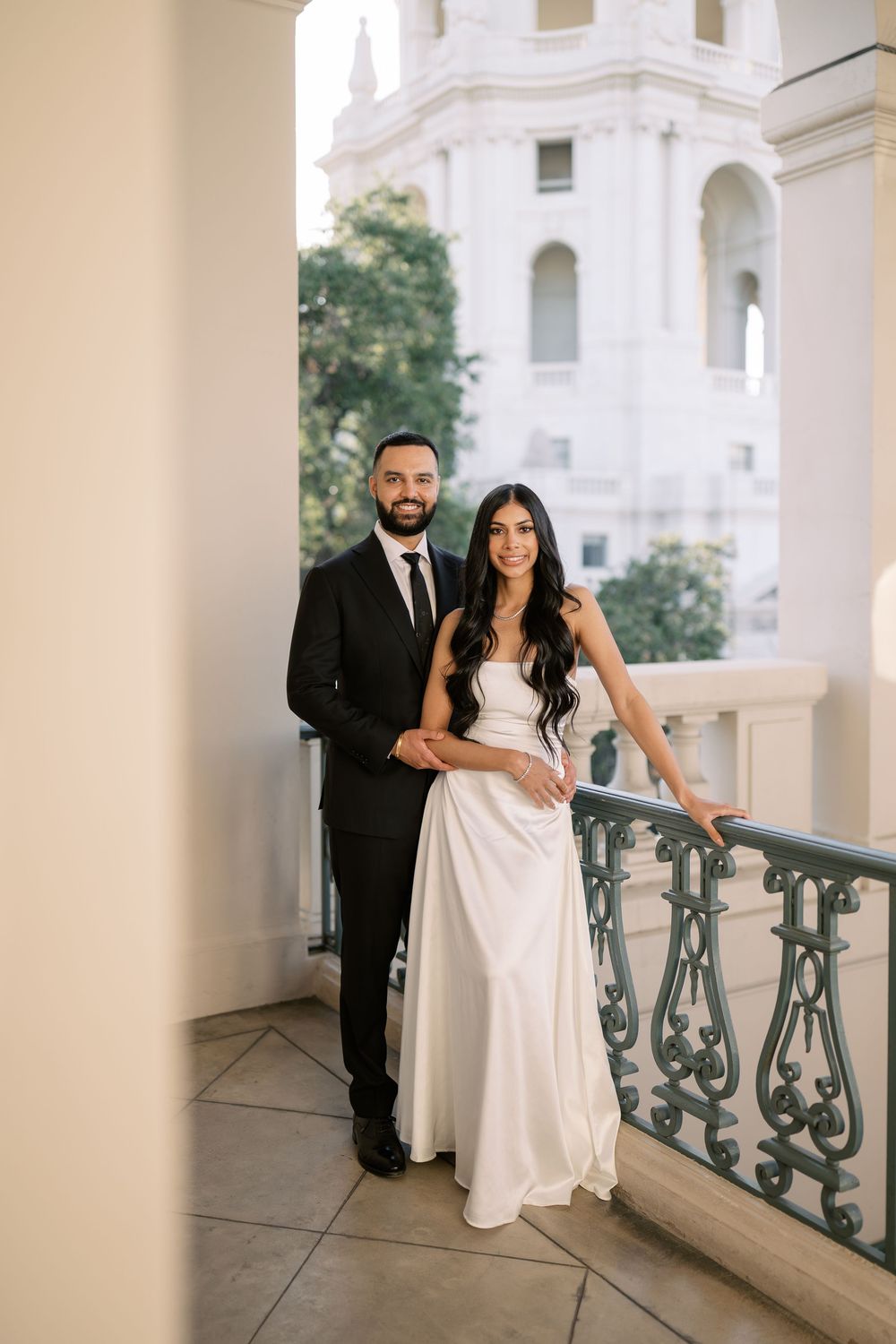 Bride and groom posing on an elegant balcony with ornate railings and a domed building backdrop.