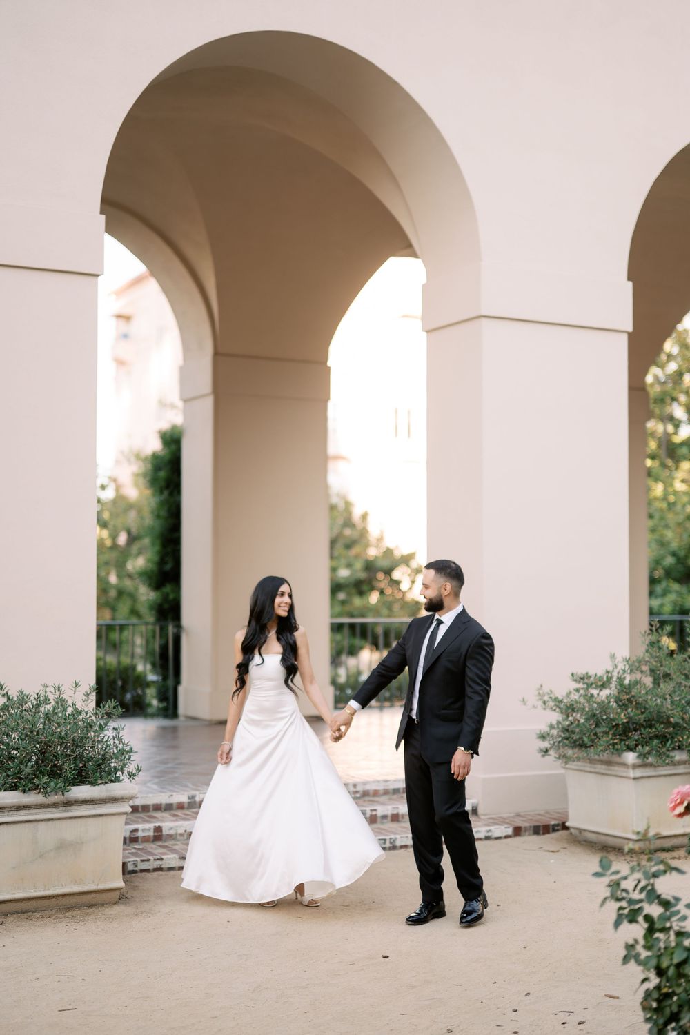 Bride and groom holding hands walking through sunlit arched colonnade with greenery.