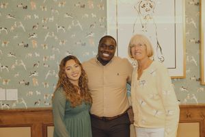 Three people smile together in a room with teal dog-themed wallpaper and framed dog illustrations.