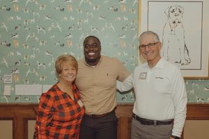Three smiling people pose together in front of a dog-patterned wallpaper at an indoor event.