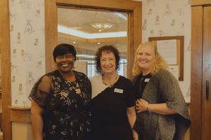 Three women smiling together at an indoor event, dressed in elegant attire against a decorative wallpapered background.