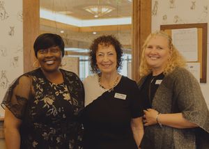 Three smiling women posing together at a formal event in an elegantly decorated room.