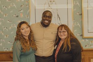 Three people pose together smiling in front of decorative bird-patterned wallpaper.