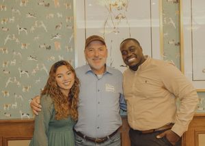 Three people smiling together indoors in front of dog-themed wallpaper and framed artwork.