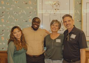 Four people smile together posing in front of dog-themed wallpaper at an indoor event.