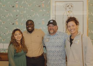 Four smiling people pose together in front of dog-themed wallpaper at a casual indoor gathering.