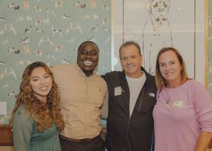 Four adults smile together in front of decorative dog-themed wallpaper at an indoor social event.