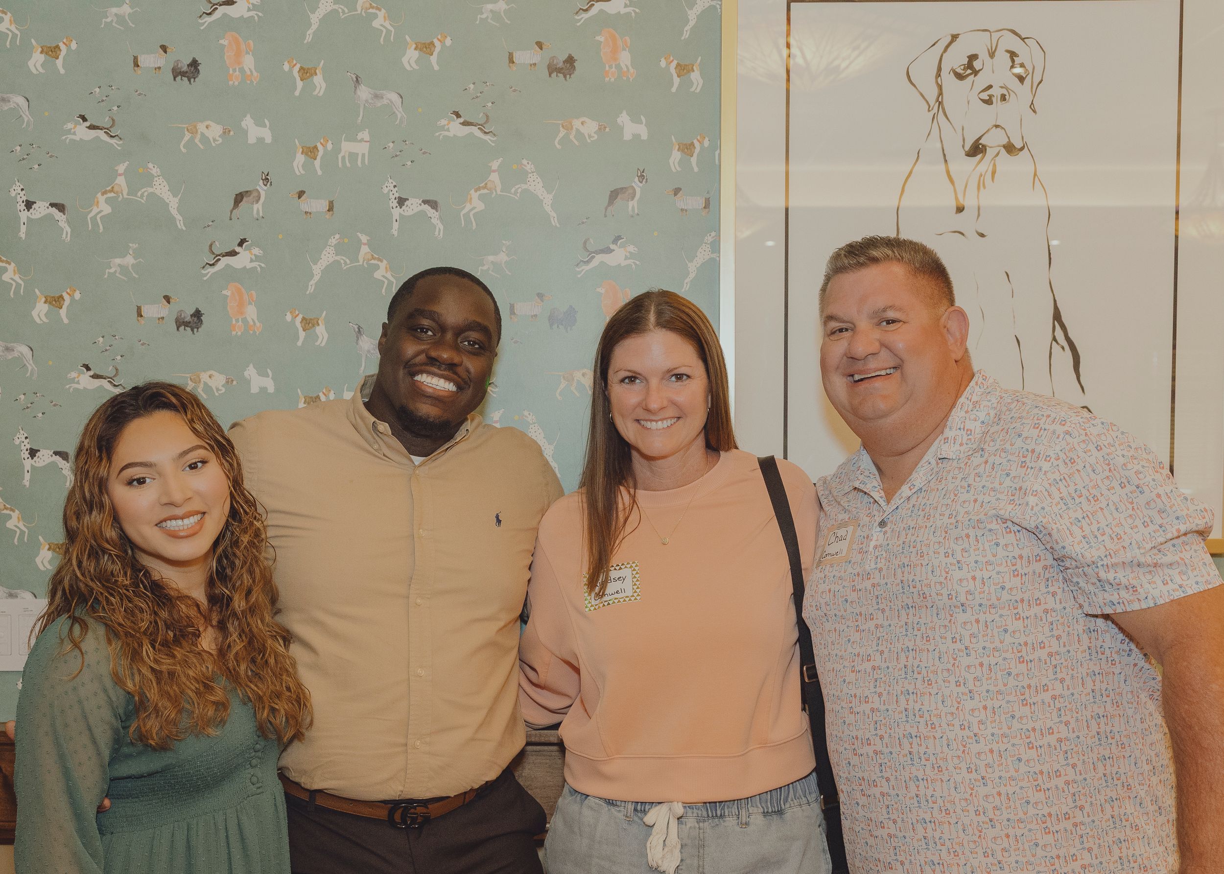 Four friends smiling together in front of decorative dog-themed wallpaper at a casual indoor gathering.