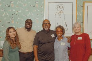 Five people smiling together indoors near a dog illustration wall art at a social gathering.