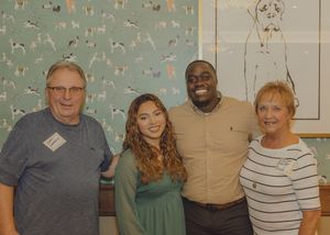 Four people smiling and posing together indoors in front of a decorative dog-themed wallpaper backdrop.