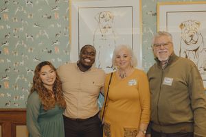 Four people smiling together in front of dog-themed wallpaper and framed dog artwork indoors.