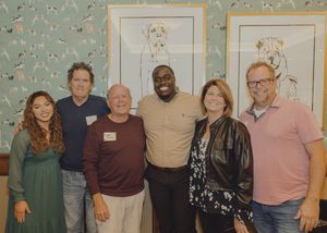 Six people pose together smiling in front of framed dog artwork at a social gathering.
