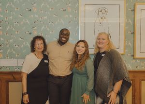 Four people smiling and posing together indoors in front of a decorative wallpaper with dog illustrations.