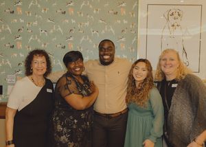 Five people smiling together in front of decorative dog-themed wallpaper at an indoor event.