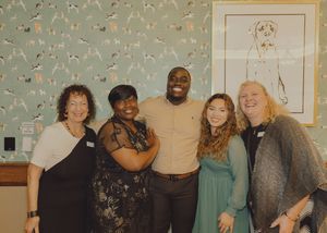 Five people smile together at an indoor gathering, standing in front of decorative dog-themed wallpaper.