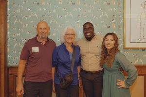 Four people smiling and posing together indoors against a decorative wallpaper backdrop at a social gathering.
