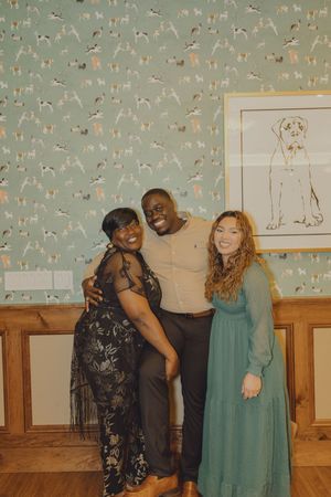 Three friends pose and smile together in a festive room with dog-themed wallpaper and framed dog artwork.