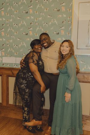 Three friends smiling and posing together at a formal event in front of green dog-print wallpaper.