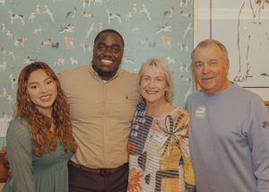 Four smiling people posing together indoors in front of a decorative wallpaper background.