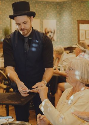 A magician in a top hat performs a card trick for a seated elderly woman at an indoor event.