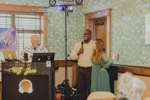 Man speaks into microphone beside woman near DJ Jose setup at event.