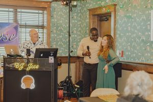 Man and woman stand together at microphone beside DJ Jose at indoor event.
