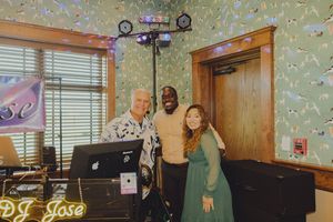 Three people pose together next to DJ Jose equipment with decorative party lights.