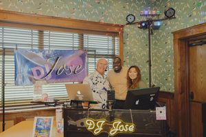 Three people pose behind a DJ Jose booth with laptops and colorful lighting at an indoor event.