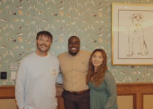 Three people posing together smiling in front of green dog-themed wallpaper with framed art.