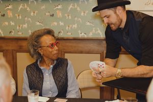 A magician in a black hat shows a card trick to a woman with red glasses at a dining table.