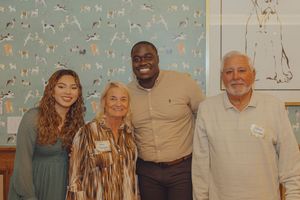Four people smiling and posing together indoors in front of a decorative wallpaper featuring animals.