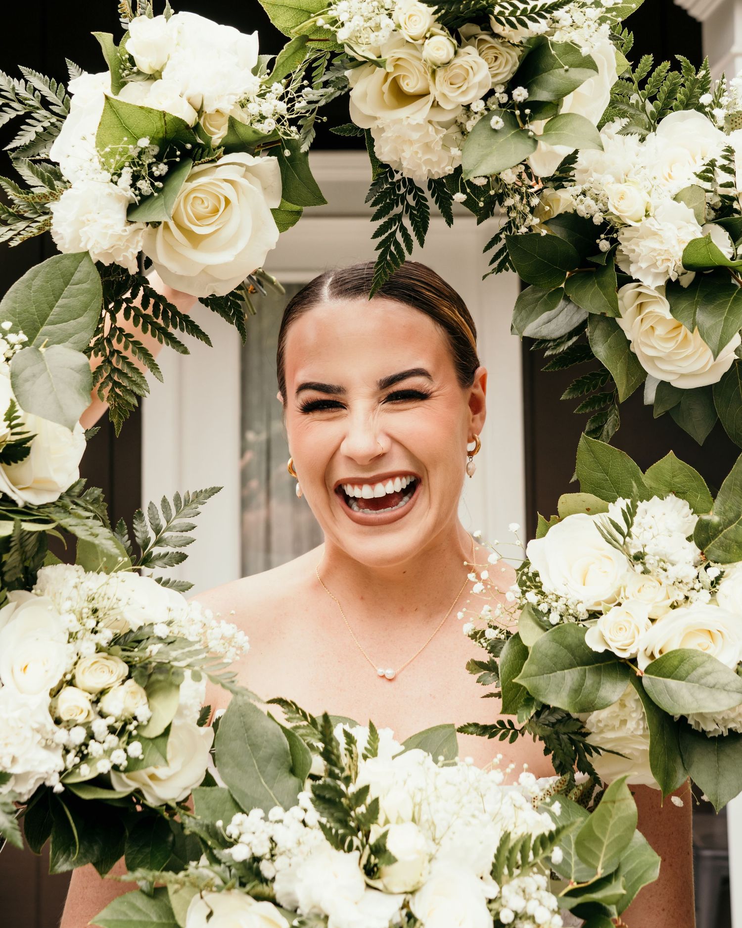 Smiling bride framed by a lush floral arch of white roses, greenery, and baby's breath on her wedding day.