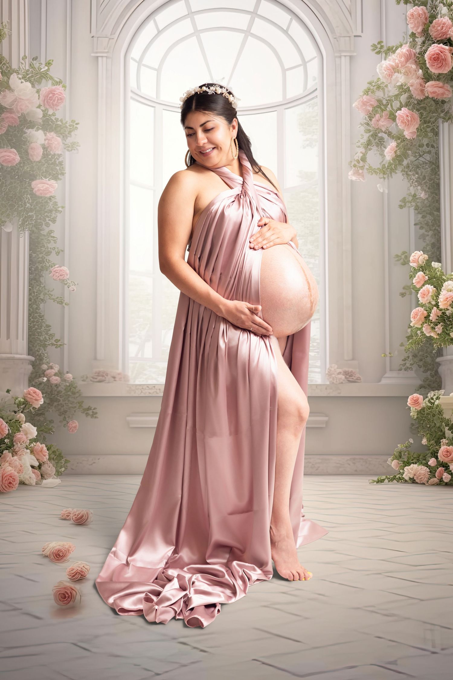 Pregnant woman in pink satin gown cradling baby bump, standing among roses in elegant white room.