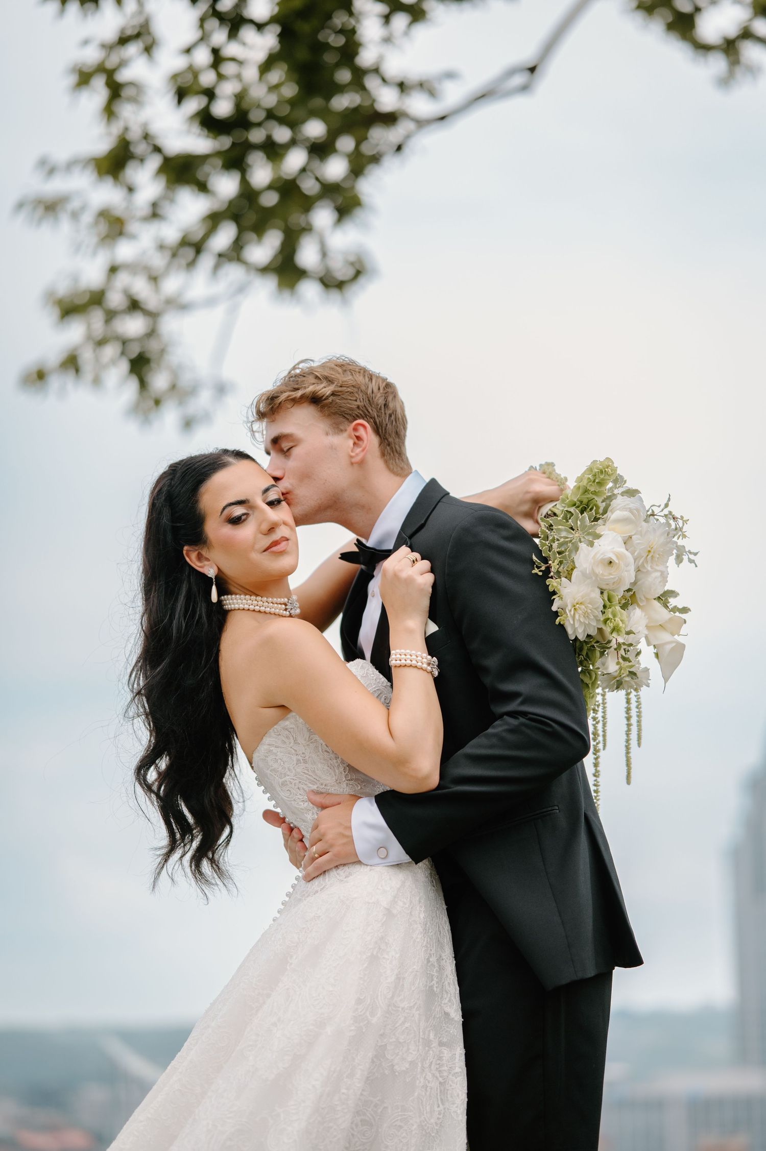 Groom in black tuxedo kisses bride in white lace gown holding white floral bouquet outdoors in Cincinnati