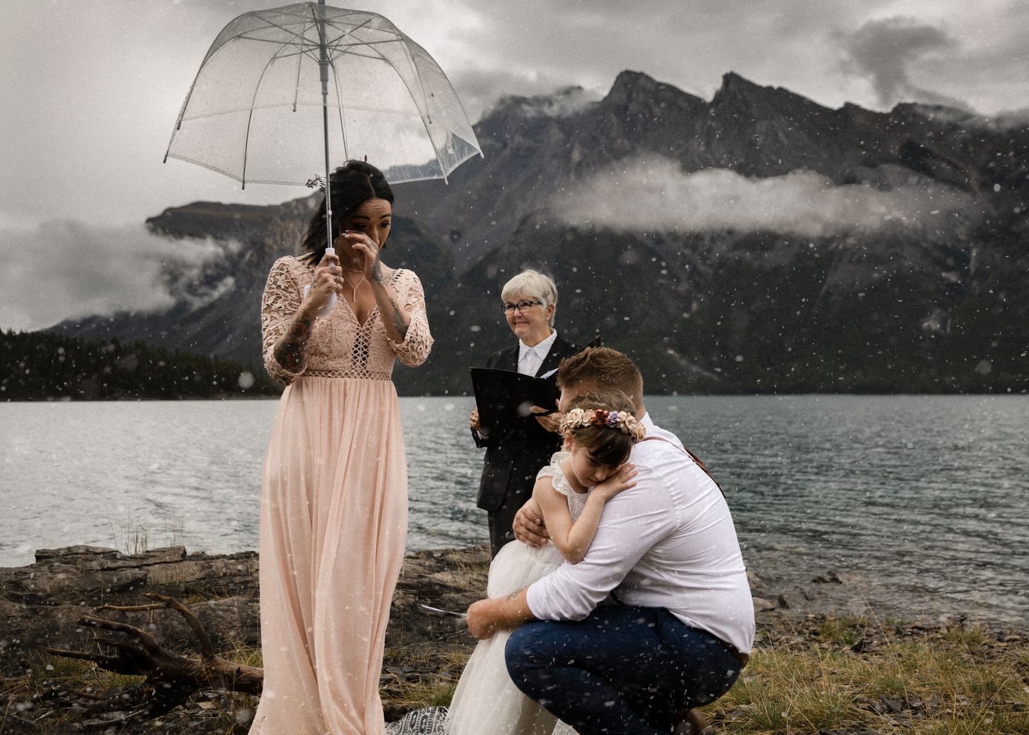Bride in blush lace wiping tears under a clear umbrella at a snowy lakeside ceremony, by a Banff wedding photographer
