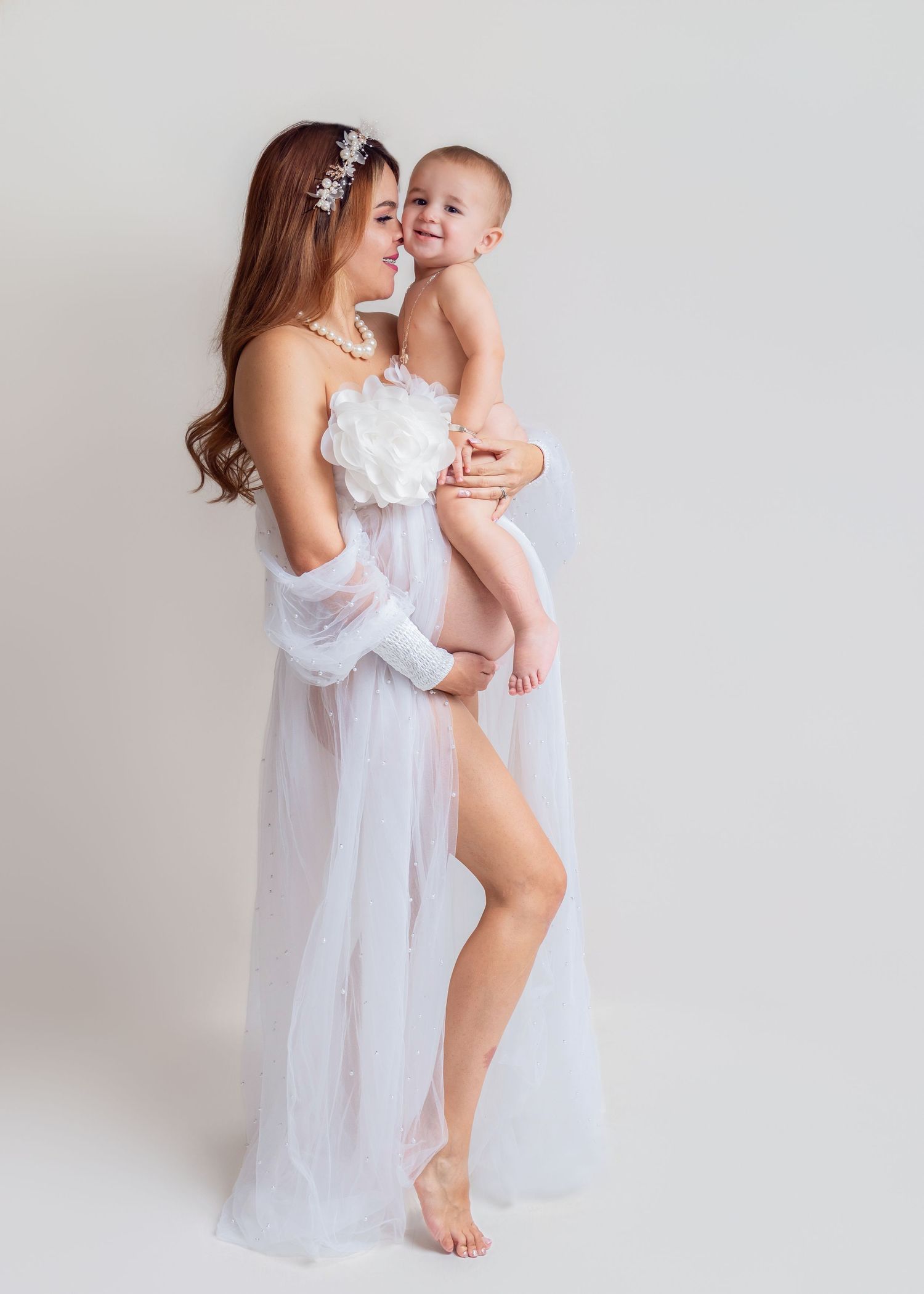 A young mother in a white sheer gown and floral headpiece holds her smiling baby in a bright studio setting.