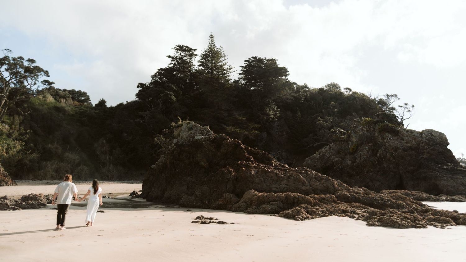 Couple holding hands walking along a sandy beach toward rocky coastal cliffs topped with lush green trees under a soft cloudy sky.
