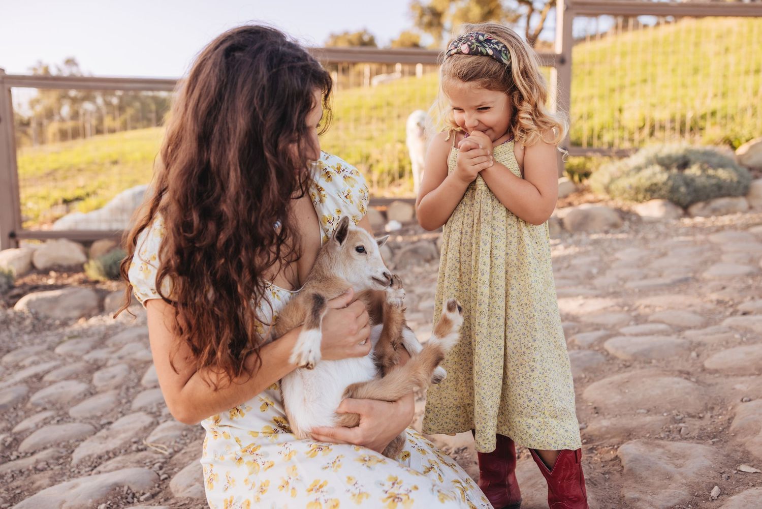 Woman in a floral dress holds a baby goat while a smiling little girl in a green dress and red boots watches at a petting farm.