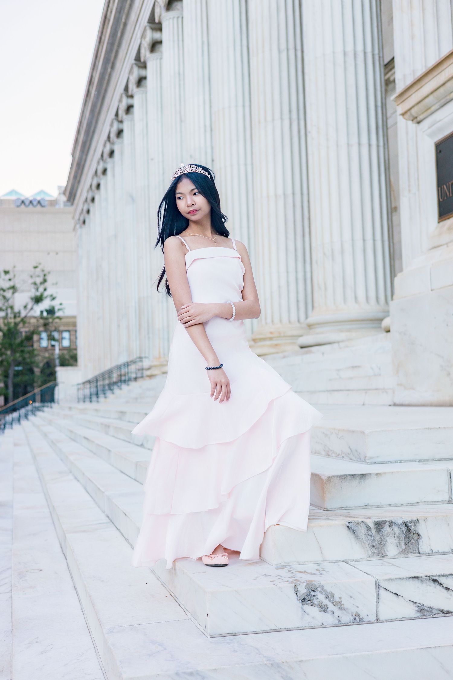 Young woman in elegant pink gown with tiara poses gracefully on marble steps of classical building.