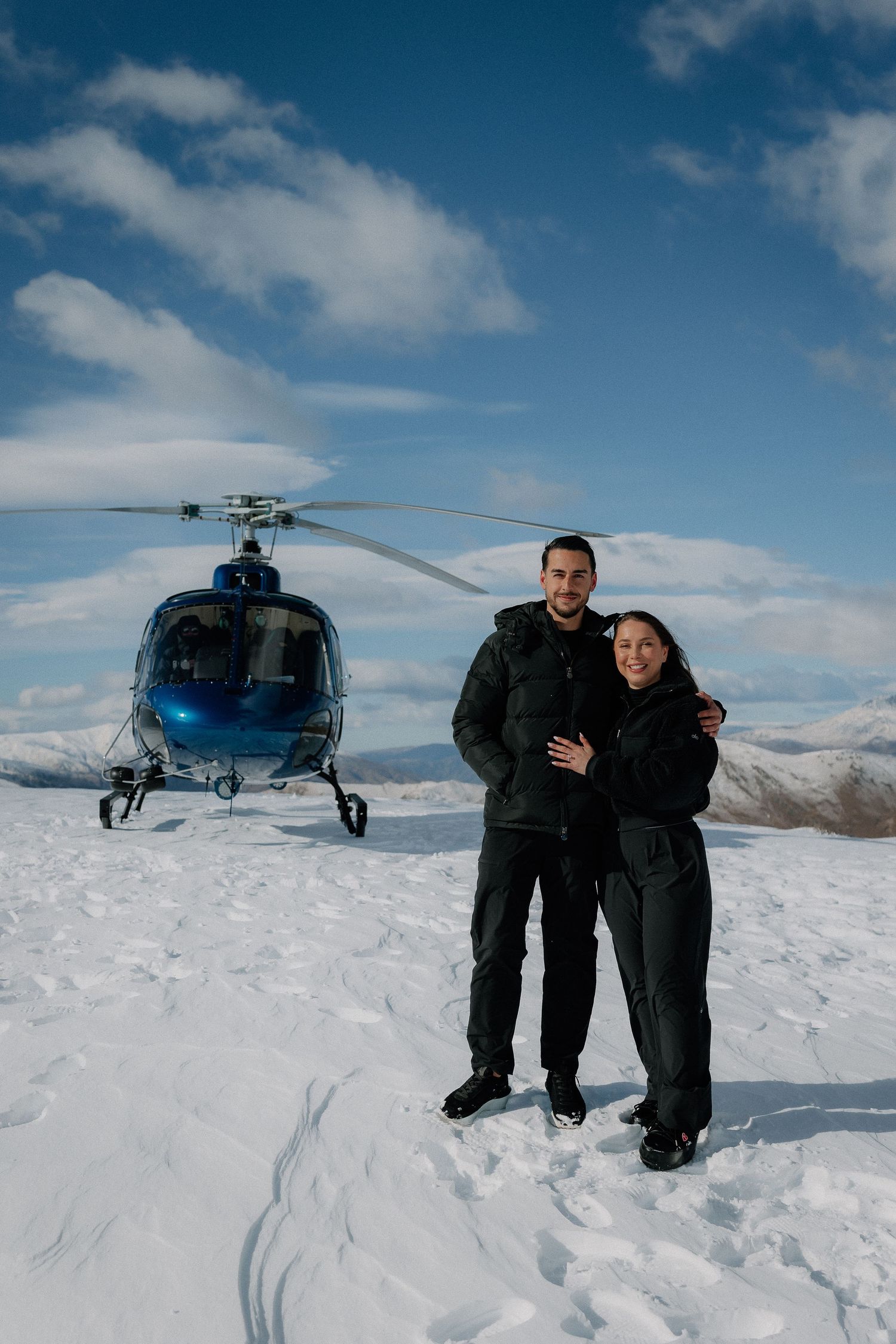 Couple posing in front of a blue helicopter on a snowy mountain summit under a partly cloudy sky.