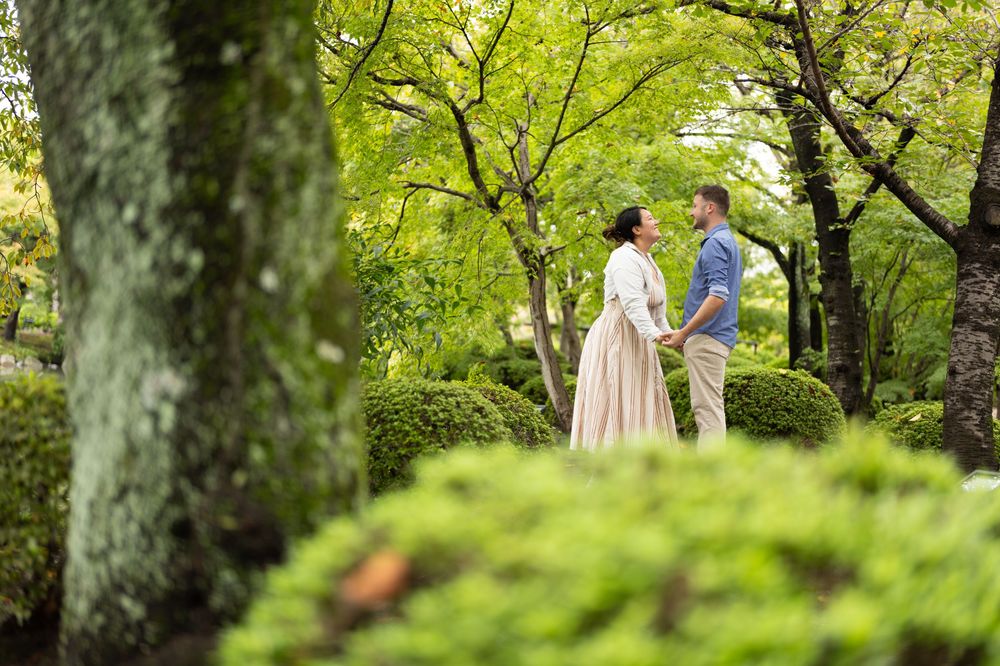 Couple in light summer outfits holding hands and facing each other in a lush green park surrounded by trees and trimmed shrubs.