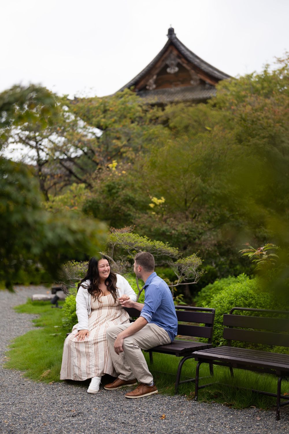 Smiling couple sitting on a wooden bench in a lush Japanese garden with a traditional temple roof rising above the green trees.