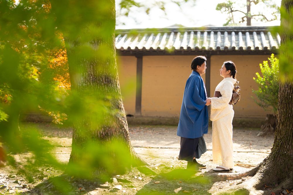 Two people in traditional-style clothing stand face to face beneath large trees in a sunlit garden near a Japanese building with a tiled roo