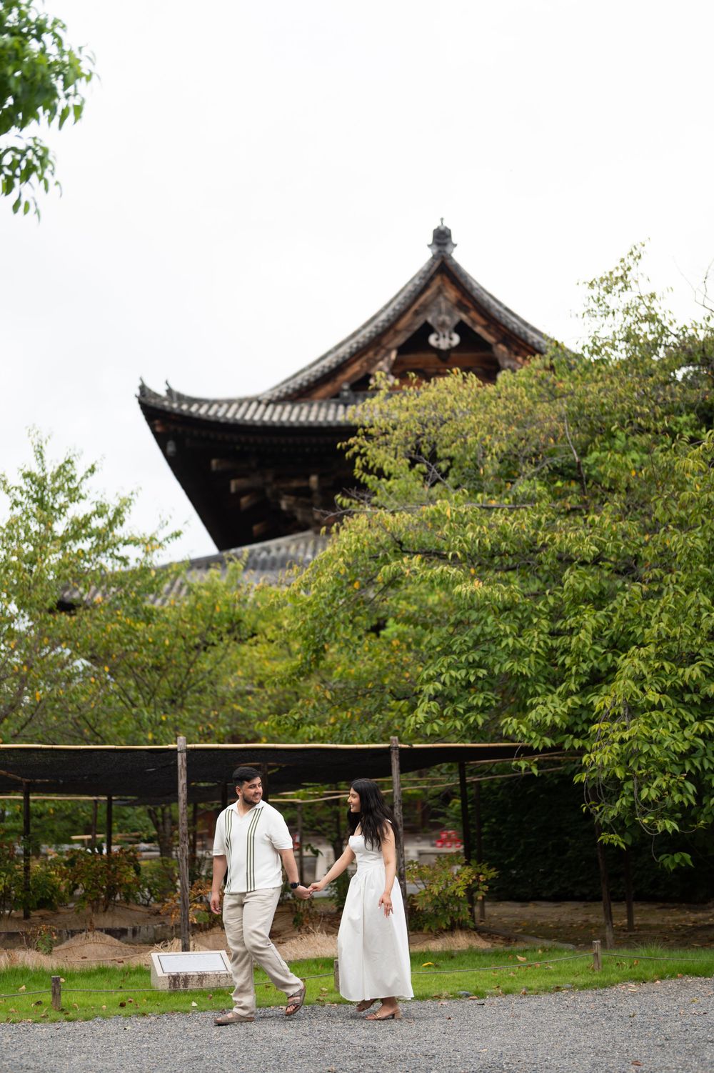 Couple holding hands and walking along a gravel path beside green trees, with a traditional Japanese temple roof rising behind them.