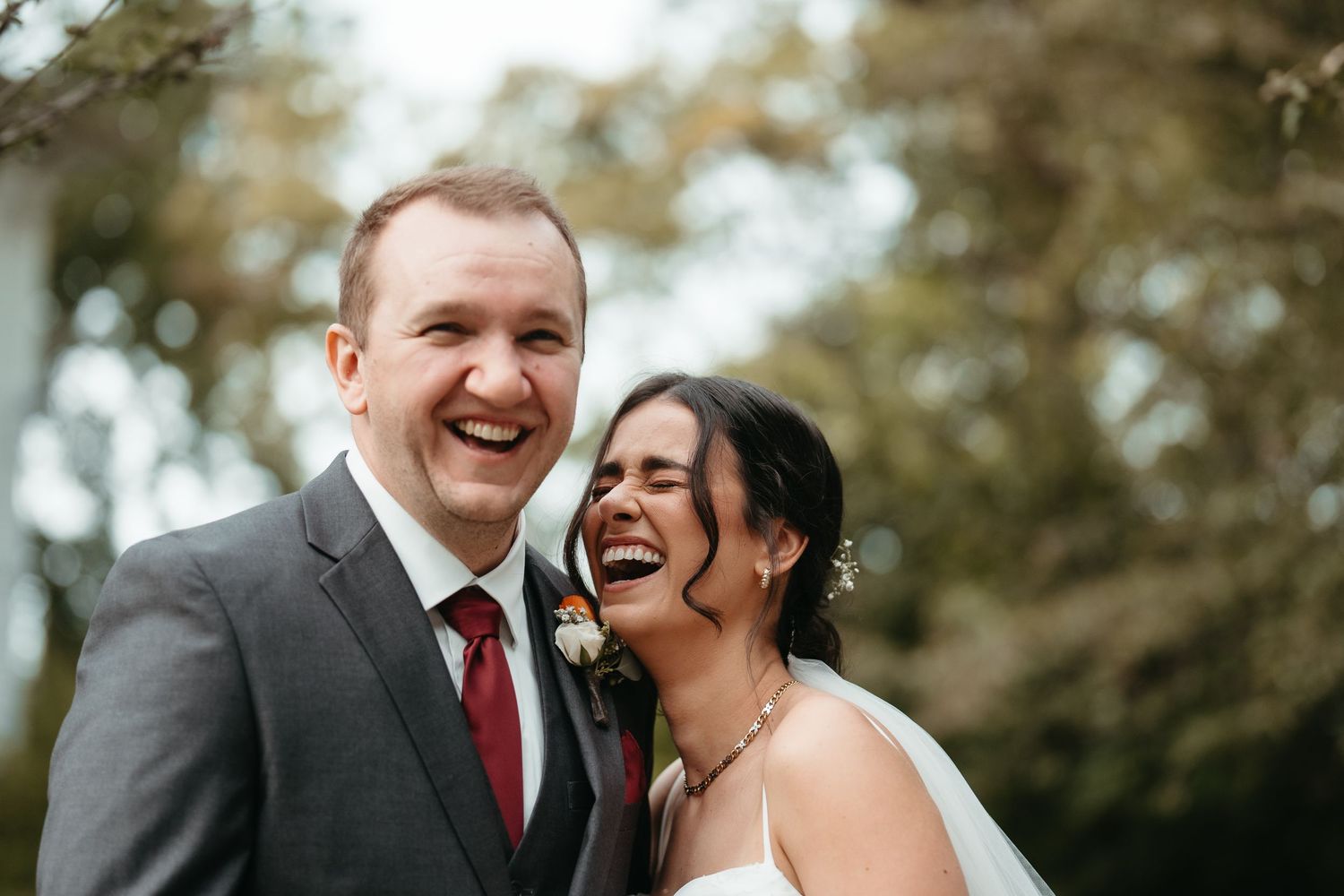 Happy bride and groom laughing together outdoors on their wedding day, groom in gray suit with red tie.