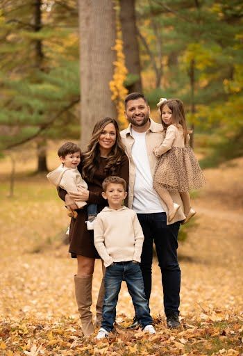 Family of five posing outdoors in autumn forest with golden leaves, two parents holding toddlers flanking older child.