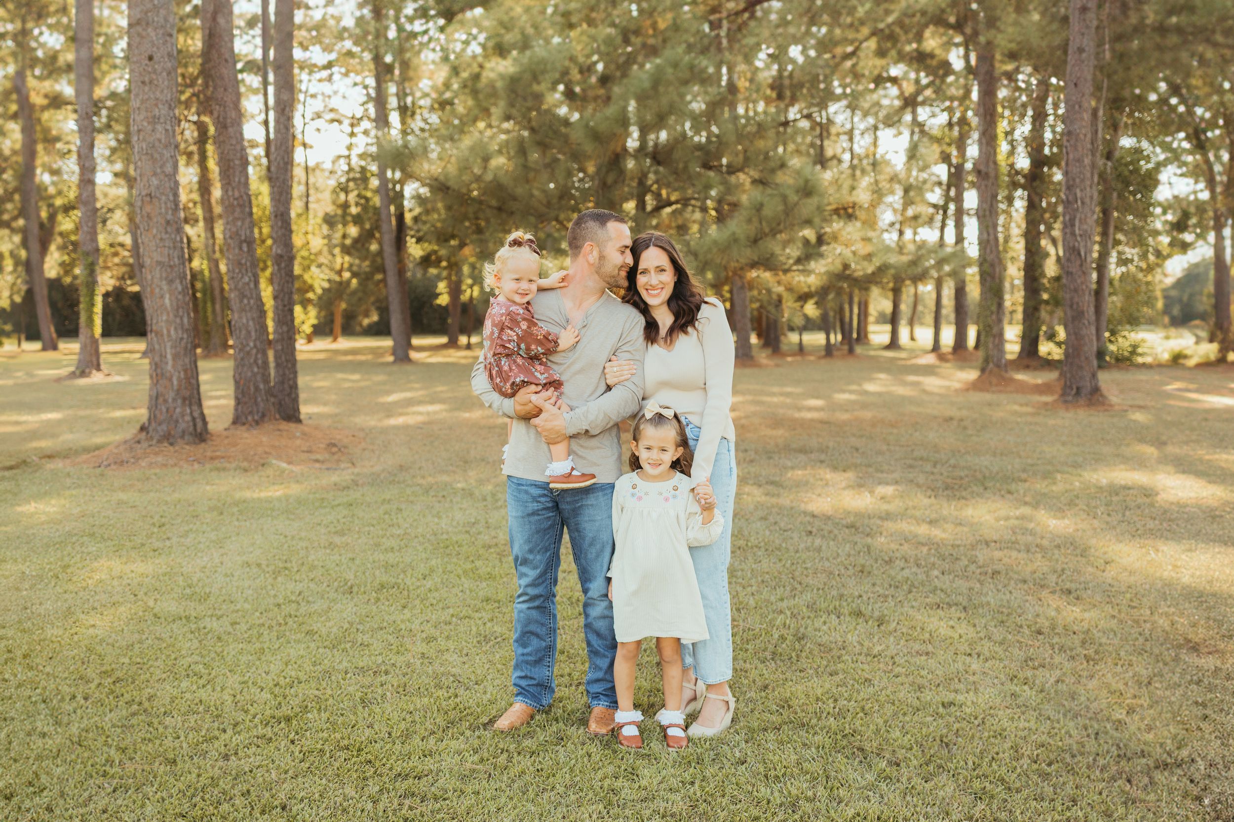 Family of four poses outdoors among tall pine trees on green grass during golden hour family portrait session.