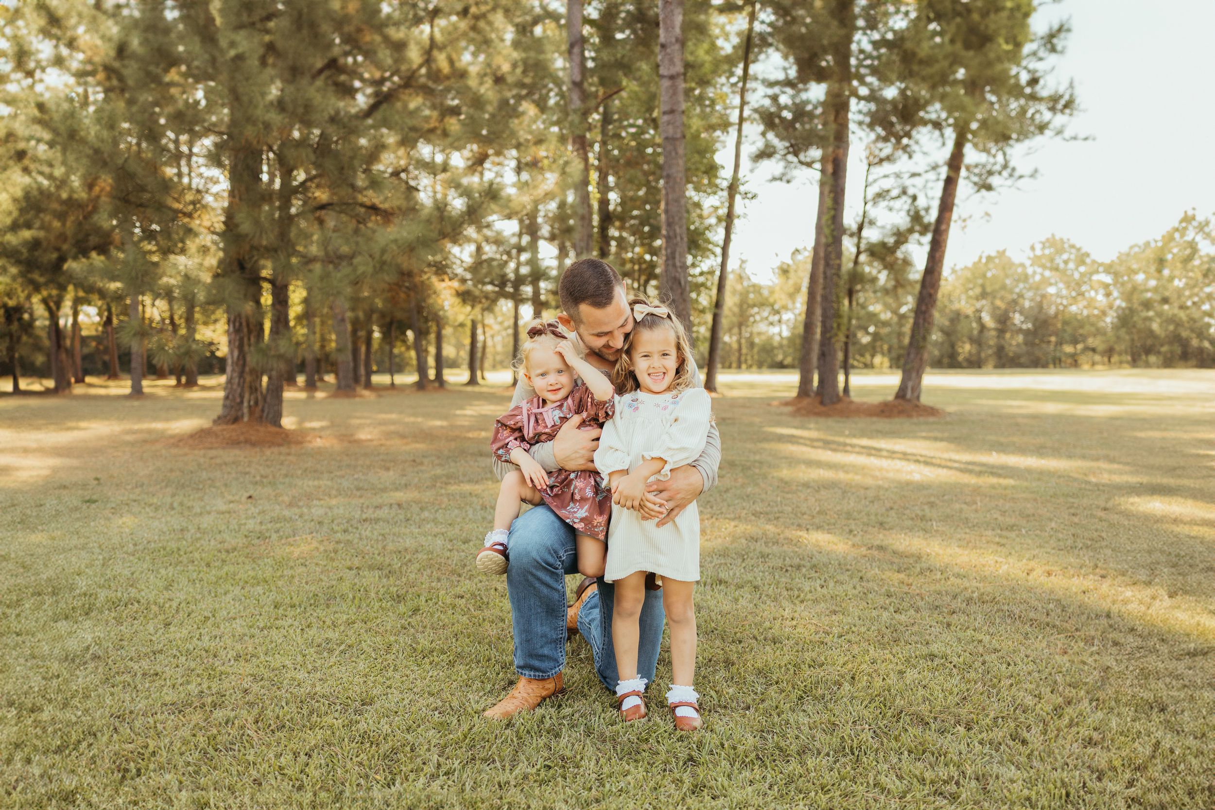 A father crouches with two young children on green grass surrounded by tall pine trees in a warm outdoor family portrait.