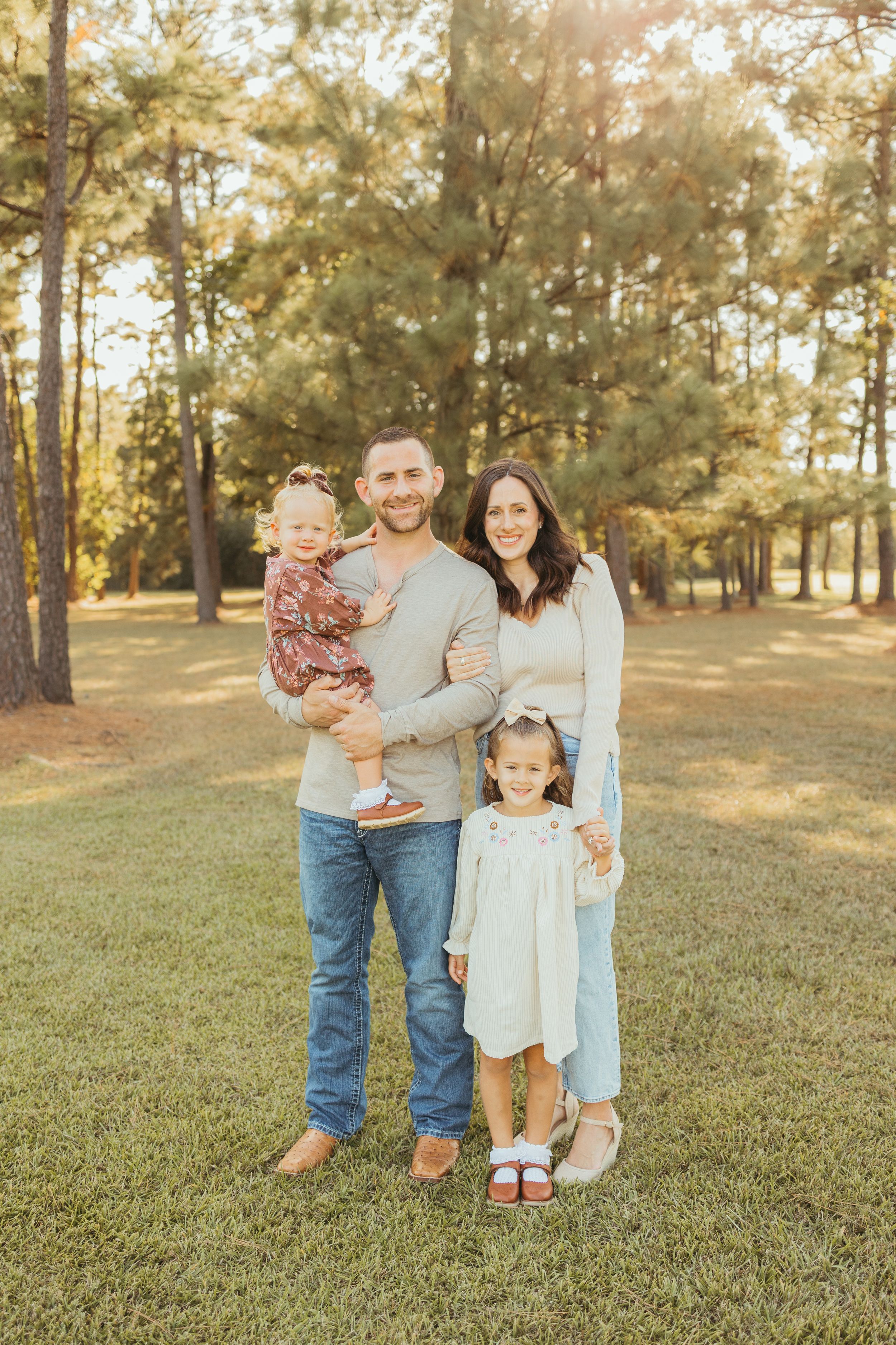 Family of four posing outdoors in a sunny pine forest, parents holding two young daughters in autumn outfits.