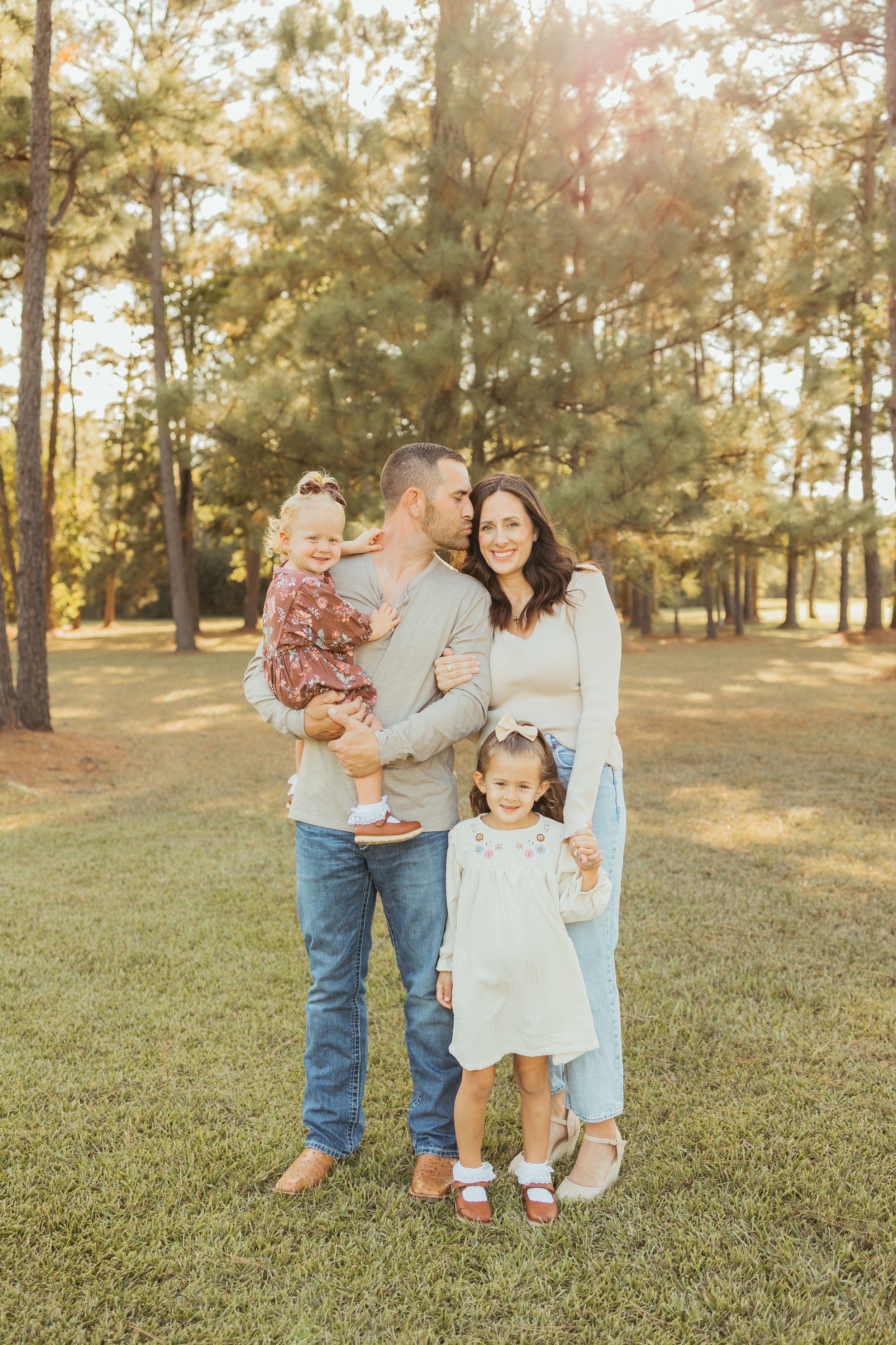 Family of four posing outdoors in a sunny pine forest, parents holding two young daughters in coordinated neutral outfits.